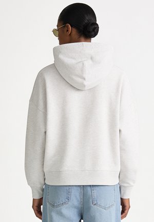 Personne aux cheveux foncés attachés en chignon, portant des lunettes de soleil, un sweat à capuche gris clair et un jean bleu clair, vue de dos sur fond blanc.