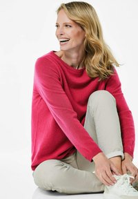 Roze trui van een zacht materiaal, met een ronde halslijn en lange mouwen, gecombineerd met lichtbeige broek en witte sneakers.