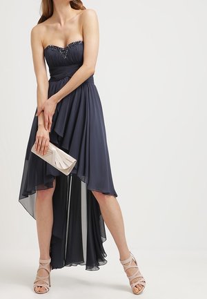 Mujer con vestido azul marino asimétrico sin tirantes con corpiño bordado, sosteniendo un bolso de mano metálico, usando tacones nude con tiras, de pie sobre fondo blanco.