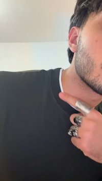 Črna majica s belim ovratnikom, vidne prstane z zapletenimi vzorci na prstih, ter kratke temne lase. Izpostavljena tekstura kože.