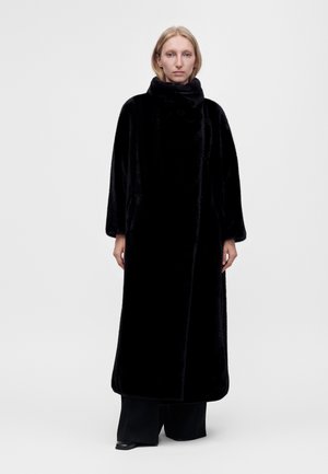 Frau mit blondem Haar trägt einen langen schwarzen Pelzmantel über schwarzen weiten Hosen und steht vor einem schlichten hellgrauen Hintergrund.