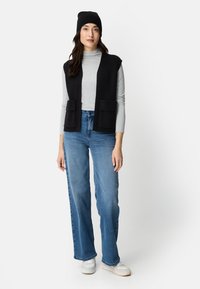 Frau trägt schwarze Mütze, schwarz ärmellose Weste mit Taschen, hellgraues Langarmshirt, blaue Jeans und weiße Turnschuhe, steht vor weißem Hintergrund.