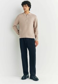 Beige långärmad tröja med krage och halvdragkedja, kombinerad med svarta byxor och svarta skor. Slät textur, avslappnad passform.