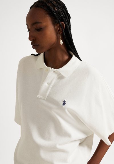 Balti polo marškinėliai, laisvo modelio, su apykakle ir trimis mygtukais. Kairėje krūtinės pusėje ryškus violetinis logotipas. Minkšta, tekstūruota medžiaga.