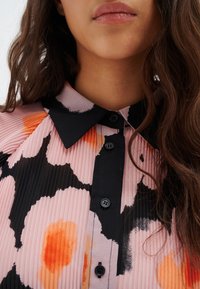 Fodrad blus med svart krage, med en rosa bas med svarta och orange abstrakta mönster, och svarta knappar längs fram.