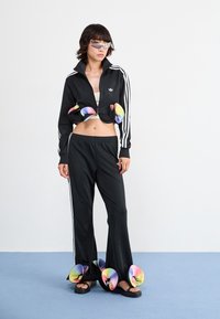 Veste de survêtement noire raccourcie avec des rayures blanches, pantalons évasés noirs et sandales avec des accents en spirale multicolores.