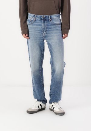Personne portant un jean bleu clair délavé à jambes droites, des baskets blanches avec des rayures noires et un haut marron à manches longues sur fond blanc.