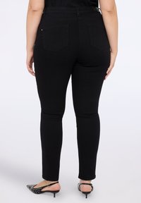 Persona con jeans negros ajustados con bolsillos traseros y tacones slingback negros con tachuelas plateadas, de pie contra un fondo blanco.