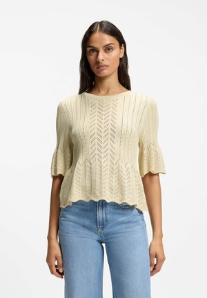 Donna che indossa un top lavorato a maglia color crema con orlo e maniche smerlati abbinato a jeans celeste a vita alta su sfondo bianco.