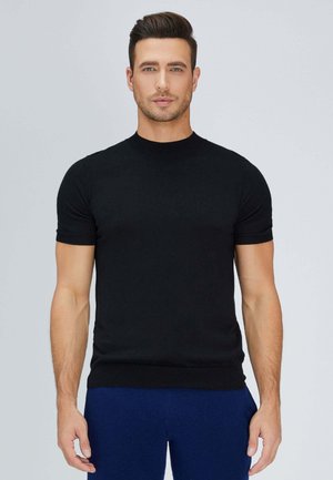 Mann mit kurzen braunen Haaren, trägt ein eng anliegendes schwarzes Kurzarmshirt und dunkelblaue Hose, steht vor einem einfarbigen hellen Hintergrund.