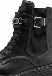Black ankle boot made of leather with a ridged elastic panel, lace-up front, and silver buckle. Features a small, decorative studded sole.