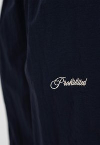 Tamsiai mėlynos spalvos audinys, ant kurio paviršiaus baltos kaligrafijos raidėmis siuvinėtas žodis „Uždrausta“, įrėmintas centre.