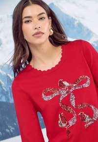 Punainen neulepaita, jossa on uurrettu kaula-aukko, joka esittelee paljetein koristellun rusetin suunnittelun ja kaksi lintomotiivia eri väreissä.