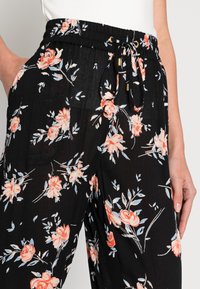 Svarta blommiga byxor med en texturerad tyg, elastisk midja, dragsko med guldtillbehör och ett mönster av persikoroser och blå löv.