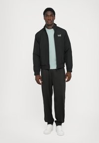 Black zip-up jacket with stand collar, paired with a light blue shirt and black trousers, all worn with white sneakers.