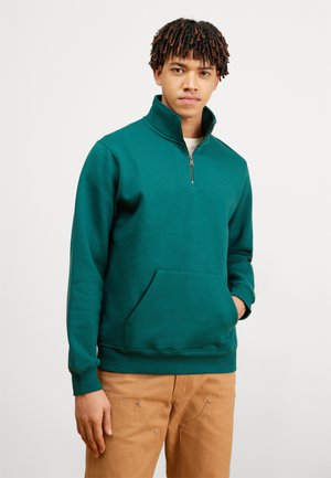 Sudadera de cuello con cremallera en color teal, fabricada en una suave mezcla de algodón, con un bolsillo canguro en la parte frontal y puños acanalados, combinada con pantalones color beige.