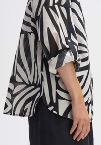 Camicia a righe bianco e nero con una vestibilità ampia, maniche arrotolate e spacco laterale, realizzata in tessuto leggero con una texture leggermente trasparente.