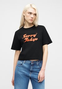 Νεαρή γυναίκα με ξανθά μαλλιά που φοράει κοντό μαύρο T-shirt με πορτοκαλί κείμενο "Sorry Tokyo" και μπλε τζιν, κοιτάζοντας προς το πλάι.