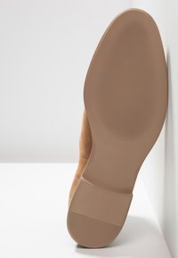 Semelle d'une chaussure en daim beige avec une semelle en caoutchouc texturée et un petit talon bloc, positionnée verticalement contre un mur blanc.
