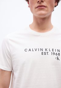 Νεαρός άνδρας που φοράει λευκό μπλουζάκι Calvin Klein με τη φράση "CALVIN KLEIN EST. 1968" και το λογότυπο "cK" τυπωμένα μπροστά.