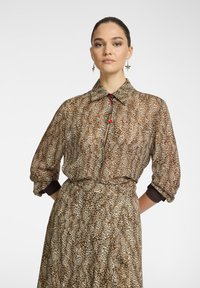 Camicia con stampa leopardata marrone e crema, con bottoni sul davanti, maniche lunghe e dettagli in bottoni rossi. Tessuto morbido e leggero con una vestibilità comoda.