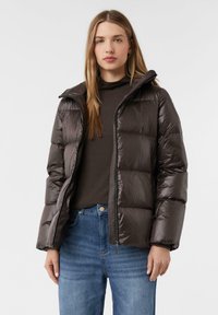 Bruine pufferjack met een glanzende, doorgestikte buitenkant, voorzien van een ritssluiting en een capuchon, gedragen over een donkere top en blauwe spijkerbroek.