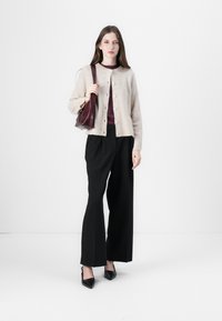Beige gebreide cardigan met ronde hals en knopen, gecombineerd met een bordeaux top, zwarte wijde broek en zwarte schoenen met puntige neuzen.