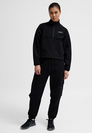 Czarny polarowy sweter z wysokim kołnierzem i zamkiem z przodu, w połączeniu z czarnymi spodniami cargo z bocznymi kieszeniami i elastycznymi mankietami. W zestawie czarne sneakersy.