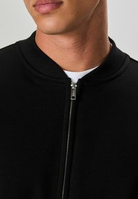 Giacca nera con zip, colletto a costine e superficie in tessuto liscio. Presenta una zip argentata sul davanti e una maglietta bianca sotto.