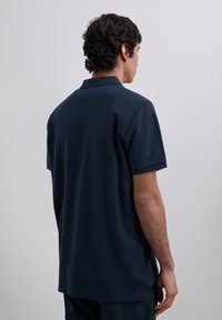 Mężczyzna z ciemnymi kręconymi włosami, ubrany w granatową koszulkę polo z krótkim rękawem, widziany od tyłu na jednolitym jasnoszarym tle.