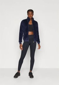 Tumesinises toonis fliisist tõmblukuga jakk, pehme tekstuuriga, kõrge krae, sobitatud mustade retuuside ja kingadega, taustal hele valge taust.