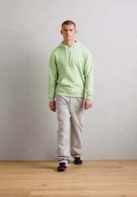 Haut à capuche vert clair avec des cordons blancs, associé à un pantalon de survêtement gris et des chaussures noires. Le mannequin se tient sur un sol en bois contre un fond neutre.