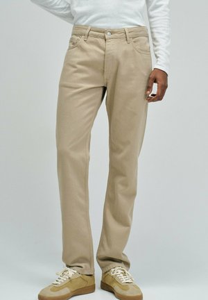 Persona con pantalones beige de pierna recta, zapatillas beige y crema, y una camiseta blanca de manga larga, de pie frente a un fondo neutro.