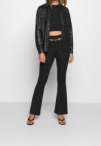 Femme portant un pantalon évasé noir avec une ceinture chaîne décorative, un crop top noir, une veste en cuir noire boutonnée et des talons ouverts noirs.