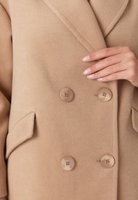 Beżowy płaszcz z wełny z teksturowanym wykończeniem, o fasonie podwójnego zapięcia, z dwoma dużymi guzikami i bocznymi kieszeniami. Zbliżenie na tkaninę i dłoń.
