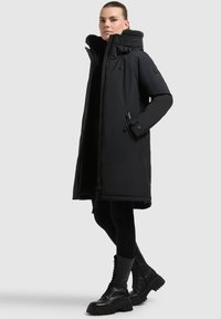 Abrigo negro largo acolchado con un cuello alto y cierre de cremallera. Presenta bolsillos laterales y un forro suave y texturizado. Combinado con botas negras gruesas.