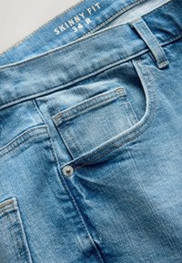 Hellblaue Jeans aus Denim mit sichtbarer Fronttasche, Gürtelschlaufe und Größenlabel "Skinny Fit 34 R" im inneren Bund.