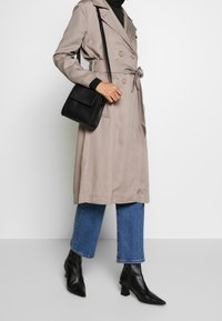Trench coat beige doppiopetto con cintura, fila singola di bottoni, abbinato a jeans blu a 3/4 e stivaletti neri con tacco.