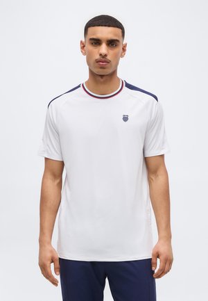 Νέος με κοντά μαλλιά που φοράει λευκό αθλητικό T-shirt με ναυτικό μπλε ρίγες στους ώμους και ένα μικρό λογότυπο στο στήθος.