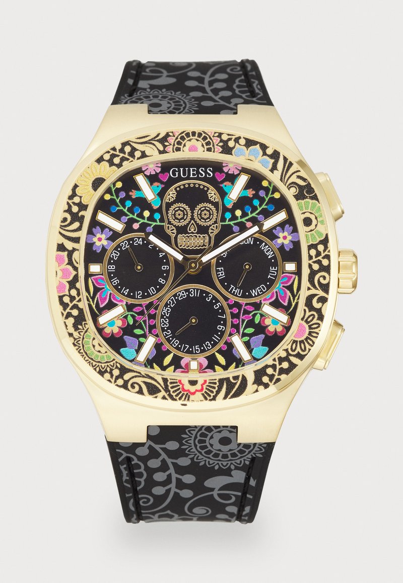 Goudkleurige chronograafhorloge met een zwarte rubberen band met patroon. Bevat een doodskopontwerp, kleurrijke bloemaccenten en een zwarte wijzerplaat met drie subdialen.