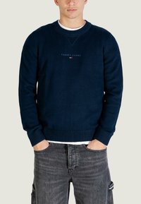 Felpa a maglia blu navy con scollo a girocollo, polsini e orlo a coste. Logo centrale in bianco e rosso. Indossata sopra una camicia bianca con jeans neri strappati.