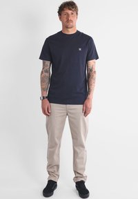 T-shirt en coton bleu marine avec un petit logo sur la poitrine, associé à un pantalon chino beige et des baskets noires ; manches courtes et col rond.