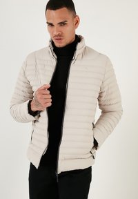 Lys beige pufferjakke med lynlås foran, høj krave og vandret quiltet design. Har sorte lynlåse og sidelommer.
