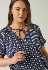 Navybluse mit weißen Punkten, kurzen Ärmeln, Bindedetail am Ausschnitt und lockerer Passform. Weicher Stoff mit einer subtilen Struktur.