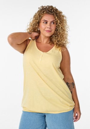 Kvinde med krøllet hår iført en gul ærmeløs top og blå jeans, poserer med den ene hånd bag nakken og ser til siden.