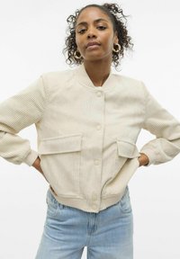 Krēmkrāsas korduroja bomberjakas ar ribotu tekstūru, priekšējiem pogām un divām krūšu kabatām. Apvienots ar gaiši ziliem džinsiem.