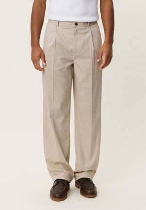 Pantalones beige plisados hechos de tela ligera. Cuentan con cierre de botón y diseño de pierna recta. Combinados con zapatos oscuros.