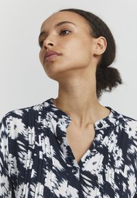 Blouse à motifs bleu marine et blanc avec un col à patte, dotée de plis et de détails en bouton, fabriquée dans un tissu léger.