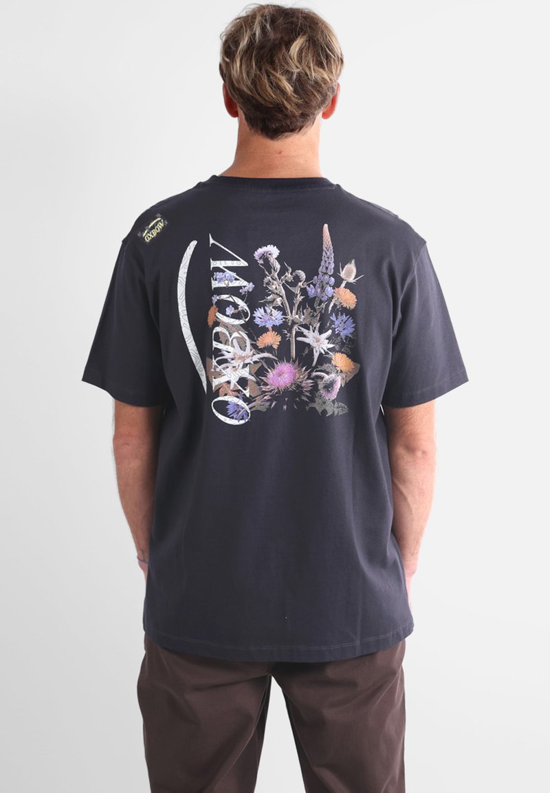 T-shirt noir en coton avec un imprimé floral coloré au dos, intégrant le texte « Rainbow » dans le design et des manches courtes.
