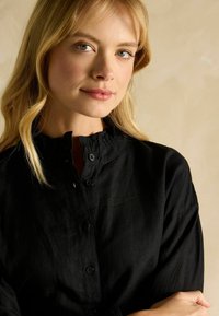 Blonde woman with light eyes wearing a black buttoned shirt with ruffled collar, standing against a beige background with arms crossed.